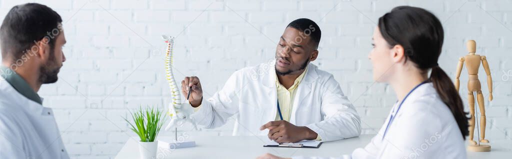 african american physician pointing at spine model during meeting with colleagues, banner