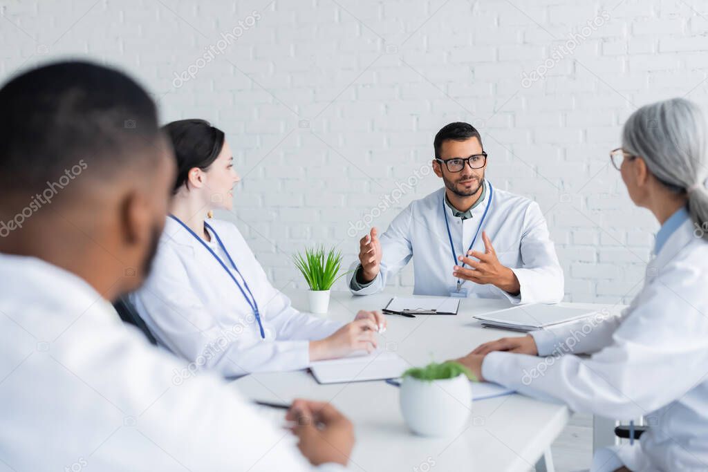 doctor in eyeglasses gesturing while talking to blurred multiethnic colleagues