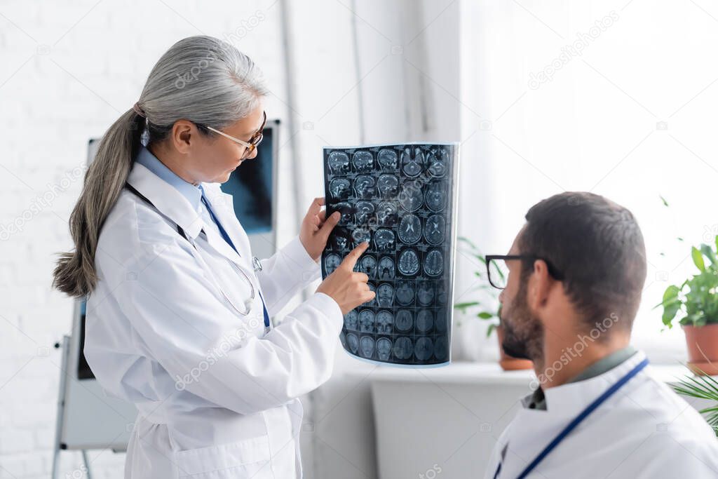 mature asian doctor pointing at head mri scan to colleague on blurred foreground