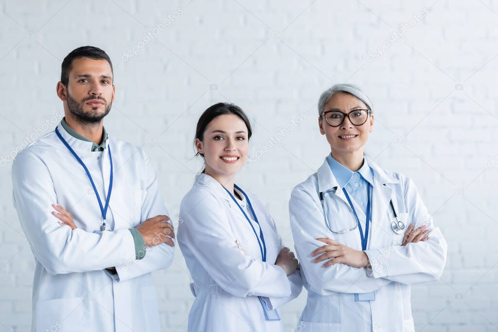 happy multiethnic doctors in white coats looking at camera while standing with crossed arms