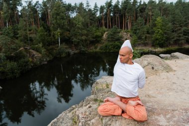 Beyaz eşofmanlı Budist Lotus pozunda meditasyon yaparken elleri arkada nehir üzerinde kayalık kayalıklarda duruyor.