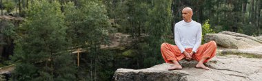Beyaz kazaklı ve harem pantolonlu Budist ormanda kayalar üzerinde meditasyon yapıyor.
