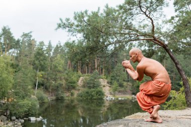 shirtless buddhist meditating in yoga pose in forest over river clipart