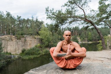 shirtless buddhist practicing extended lotus pose with praying hands on rocky cliff over river clipart