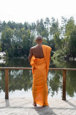 Turuncu kasaya giymiş Budist ormandaki göl kenarında meditasyon yaparken.