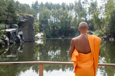 Turuncu kasaya giymiş Budist 'in orman gölü yakınlarında meditasyon yapışının arkası. 