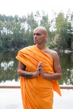 buddhist monk in traditional robe looking away while meditating near lake in forest clipart