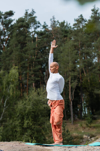buddhist in harem pants and sweatshirt practicing yoga with raised hand outdoors