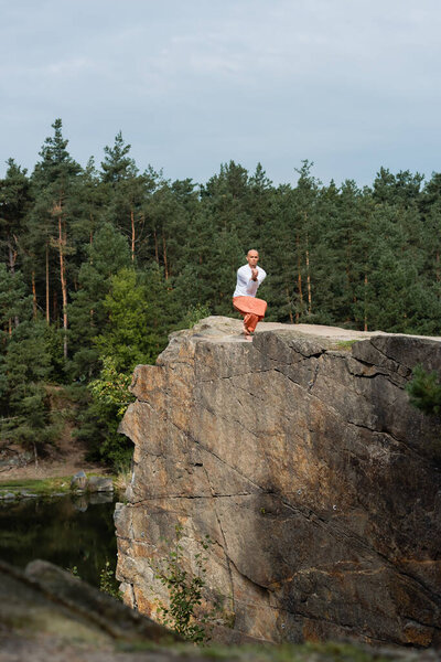 buddhist meditating in yoga pose on high rocky cliff over river in forest