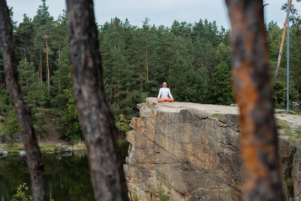 buddhist meditating in lotus pose on rocky cliff over river, blurred foreground