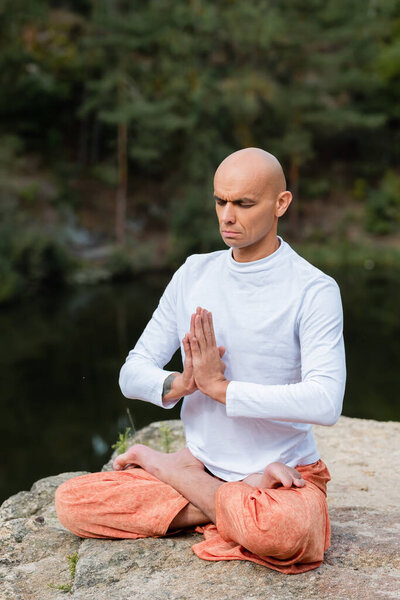 buddhist with closed eyes and praying hands meditating in lotus pose outdoors