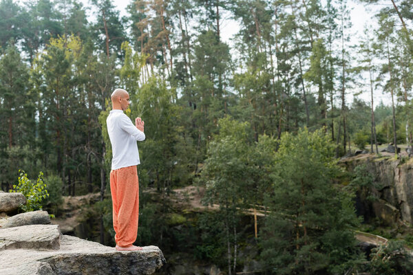 full length view of buddhist in white sweatshirt and harem pants meditating with praying hands on rocky cliff 