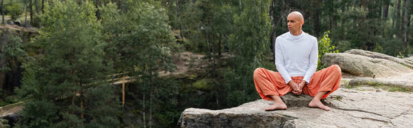 buddhist in white sweatshirt and harem pants meditating on rock in forest, banner