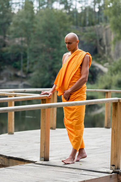 full length view of buddhist monk in orange robe walking on wooden platform in forest