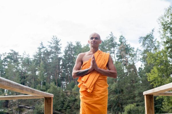 low angle view of buddhist monk meditating with praying hands near wooden fence in forest