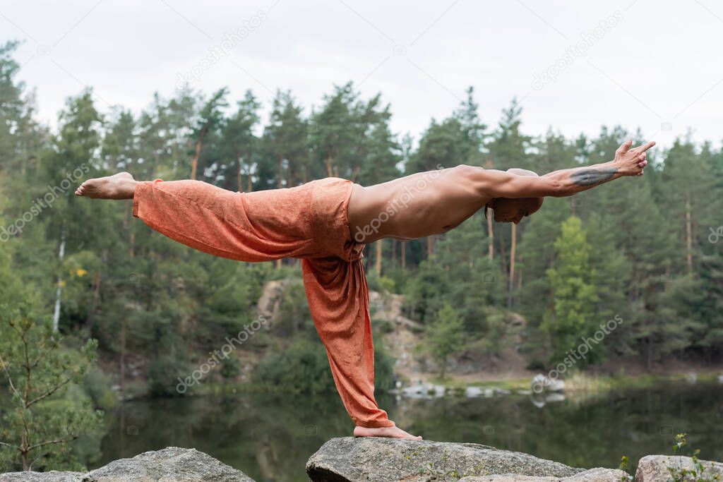 side view of shirtless buddhist meditating in half moon pose on rocks over river