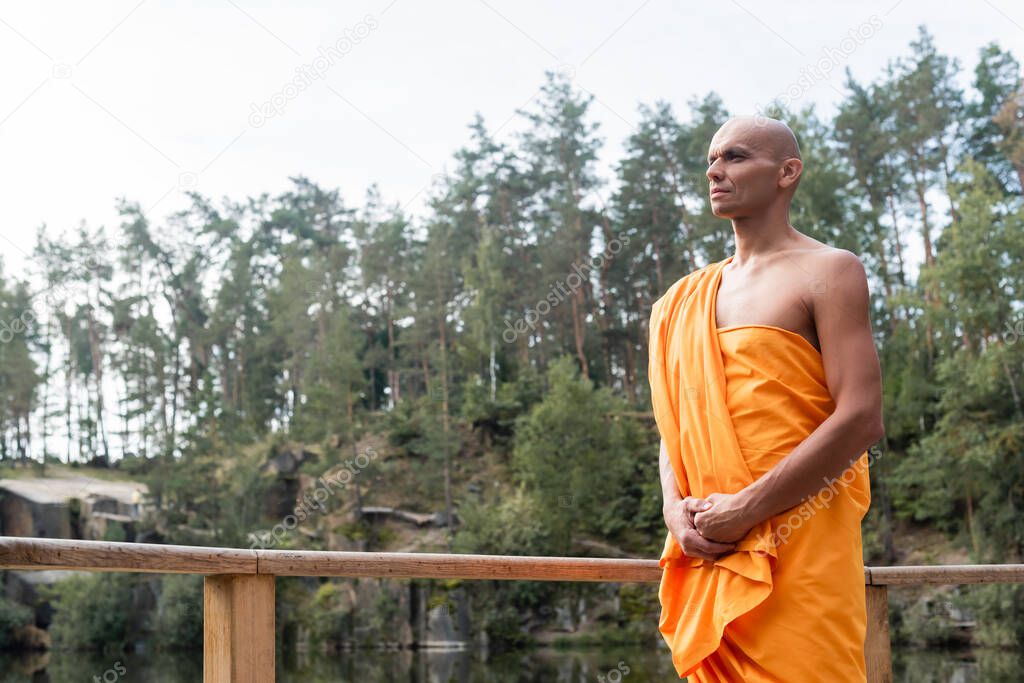 buddhist monk in orange kasaya looking away while meditating near wooden fence in forest