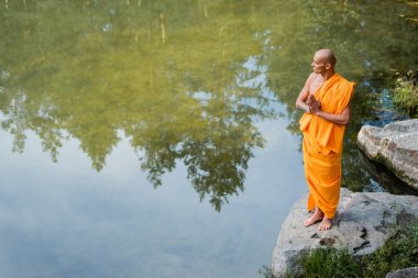 high angle view of buddhist in orange kasaya meditating with praying hands near lake clipart