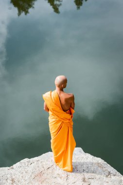 Turuncu kasaya giymiş Budist 'in gölün üzerindeki kayalık kayalıklarda meditasyon yaparken yüksek açılı görüntüsü. 