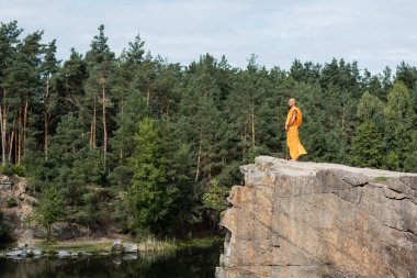 full length view of buddhist in orange robe meditating on rocky cliff over river clipart