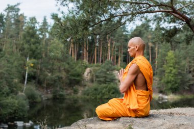 buddhist monk in orange kasaya meditating in lotus pose with praying hands near forest lake clipart