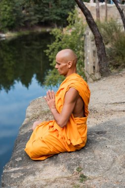 Turuncu cüppeli Budist Lotus 'ta meditasyon yapıyor. Elleri suyun yanındaki kayalık kayalıklarda dua ediyor.