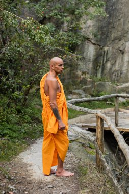 full length view of barefoot buddhist in orange robe standing near wooden fence in forest clipart