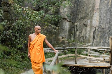 man in traditional buddhist robe walking near wooden fence in forest clipart