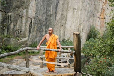 buddhist in orange kasaya standing on wooden walkway near rocks and looking away clipart