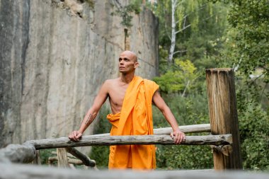 Geleneksel Budist cüppeli adam ormandaki kayaların ve ahşap çitlerin yanından uzağa bakıyor.
