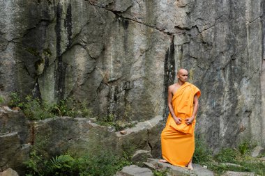 full length view of barefoot buddhist in orange robe standing near rock clipart
