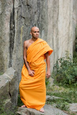 Geleneksel Budist elbiseli bir adam kayanın yanında dikilirken gözlerini kaçırıyor.