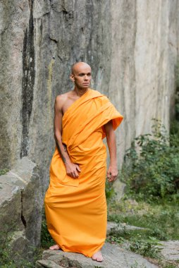 man in traditional buddhist clothes looking away while standing near rock clipart