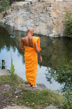 back view of barefoot buddhist in orange robe meditating near lake in forest clipart