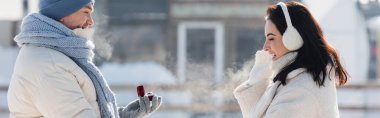 side view of man in winter hat holding box with wedding ring near happy young woman in ear muffs, banner clipart