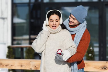 Kış şapkalı gülümseyen adam, elinde alyansla kulak manşetli genç bir kadının yanında. 