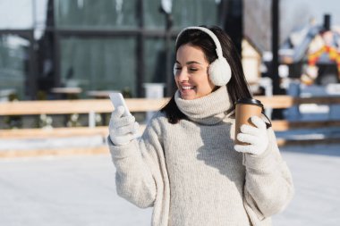 Süveter ve kulaklık takan, elinde kağıt bardak tutan ve buz pateni pistinde akıllı telefon kullanan mutlu genç bir kadın. 
