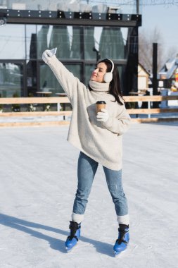 Süveter ve kulaklık takan, elinde kağıt bardak tutan ve buz pateni pistinde selfie çeken mutlu bir kadın. 