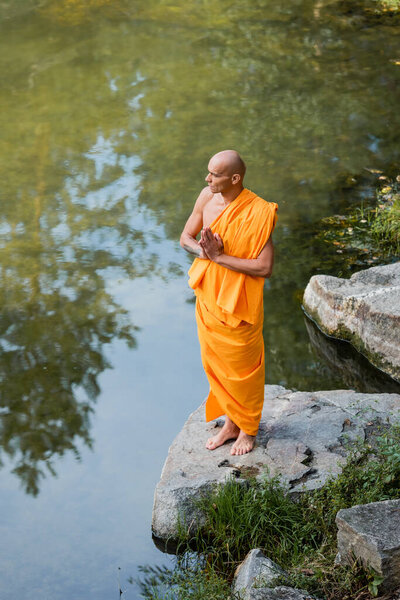 high angle view of barefoot buddhist monk in orange robe praying near river