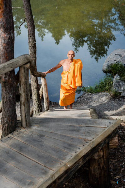 высокий угол зрения буддистов в оранжевом халате, идущих по деревянной лестнице возле озера
