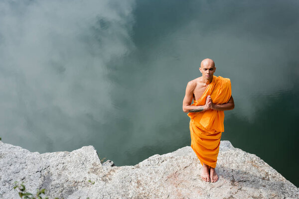 high angle view of buddhist in orange robe praying on rocky cliff over water