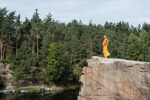 full length view of buddhist in orange robe meditating on rocky cliff over river