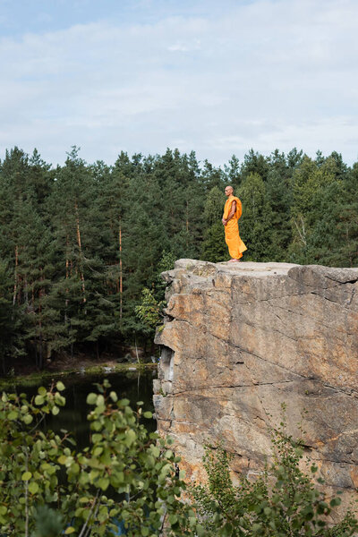 side view of buddhist in orange kasaya meditating on high rocky cliff over lake in forest