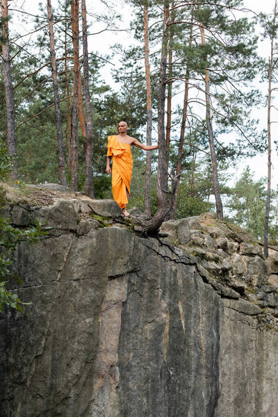 full length view of buddhist monk in orange kasaya walking on rocky cliff in forest
