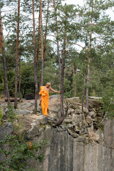ful length view of buddhist in orange robe walking on rocky cliff in forest