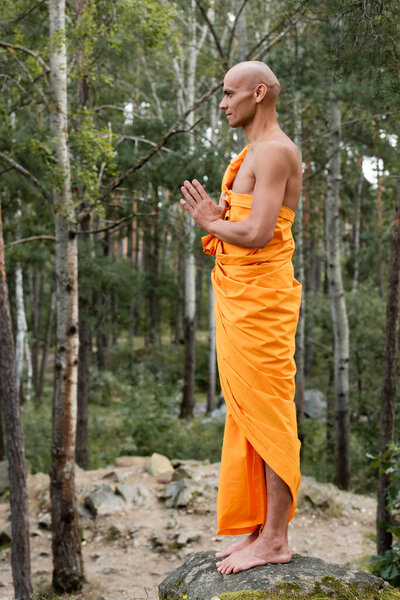 side view of barefoot buddhist in orange kasaya praying in forest