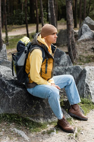 旅行者はバックパックを着て暖かいベストとビーニーを着て森の石の上に座って — ストック写真