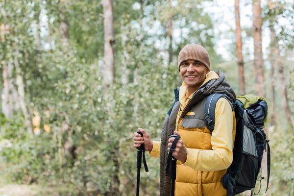 cheerful tourist with backpack and trekking poles looking away in forest