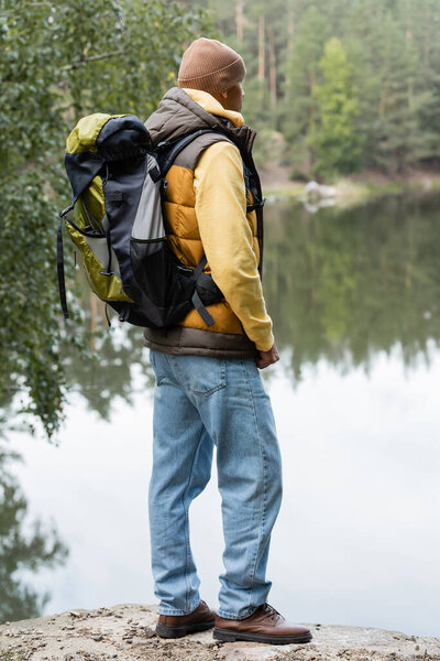 турист в теплом западе и джинсы глядя на озеро в осеннем лесу