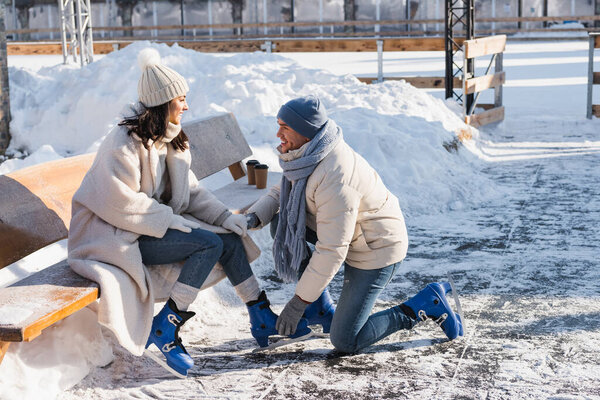 happy man wearing ice skates on cheerful girlfriend in winter hat